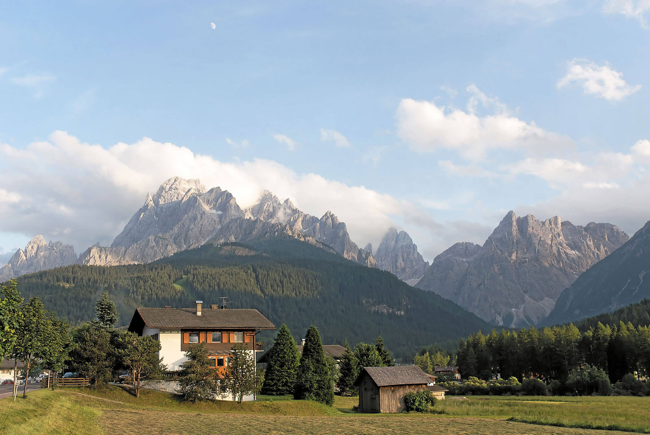Ein Haus und kleine Gebäude stehen in einem grasbewachsenen Feld mit hohen, felsigen Bergen im Hintergrund unter einem blauen Himmel. - Hotel Mooserhof