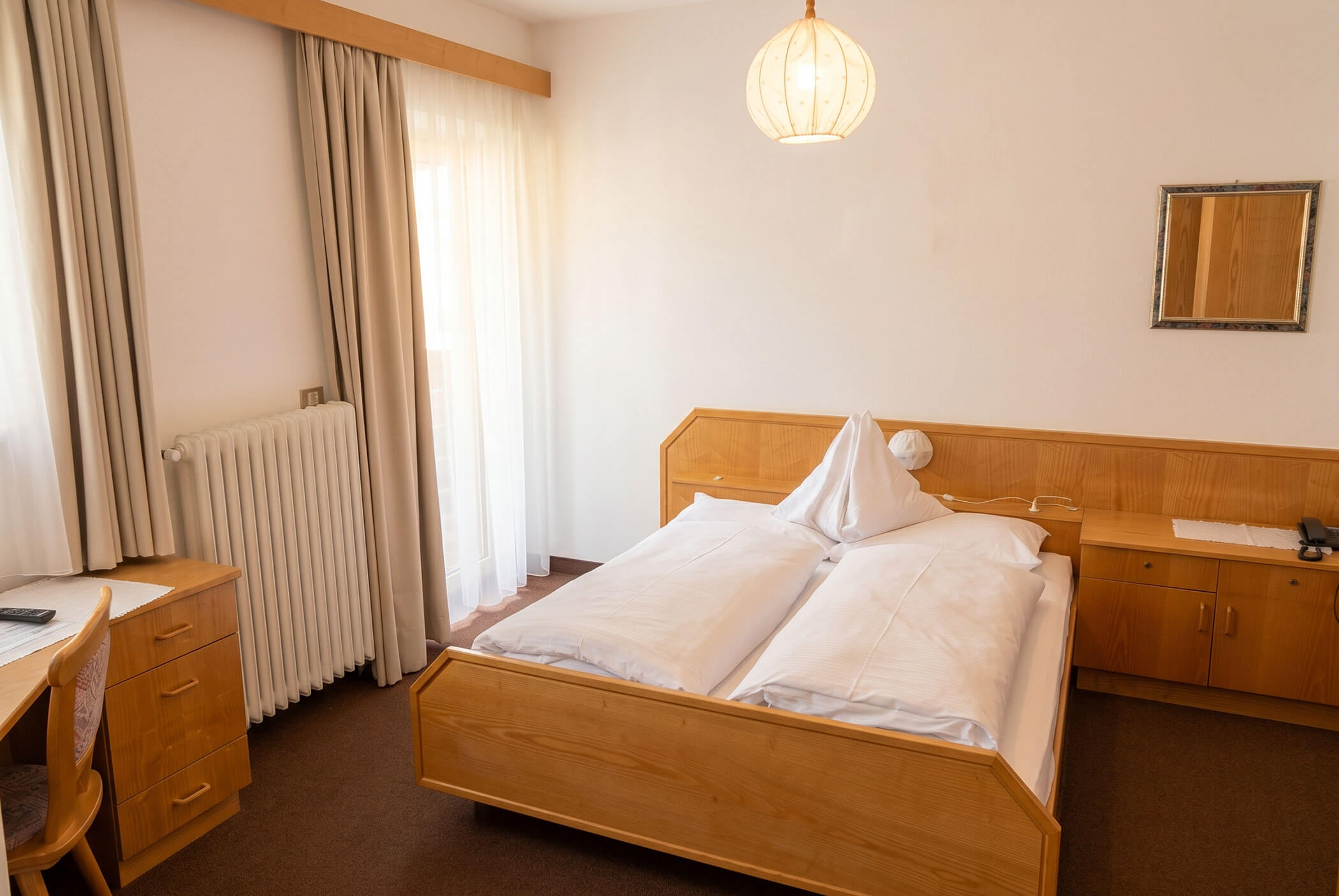 Ein ordentlich gemachtes Hotelbett mit hellen Holzmöbeln und ein Fenster mit beigen Vorhängen in einem hellen Zimmer. - Hotel Mooserhof