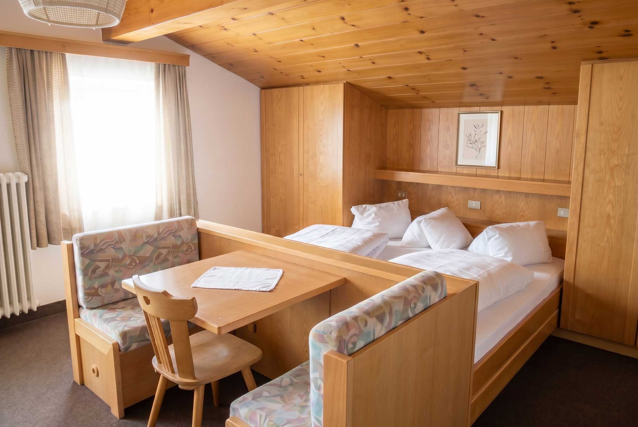 Ein gemütliches Holzzimmer mit einem Bett, einem Tisch, Stühlen, einer Sitzbank und einem Fenster mit Vorhängen. - Hotel Mooserhof