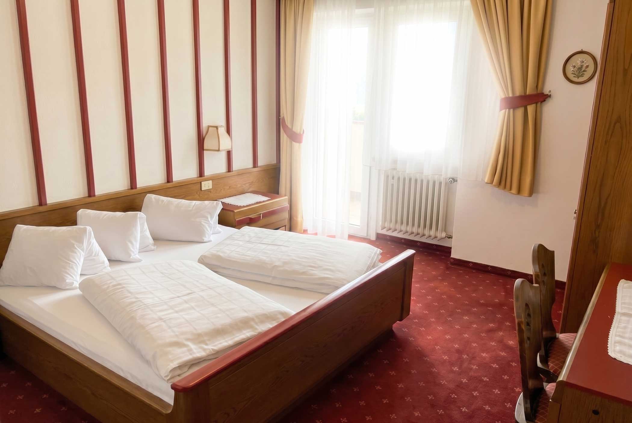 Gepflegtes Hotelzimmer mit einem Doppelbett, rotem Teppich, Holzmöbeln und Sonnenlicht durch transparente Vorhänge. - Hotel Mooserhof