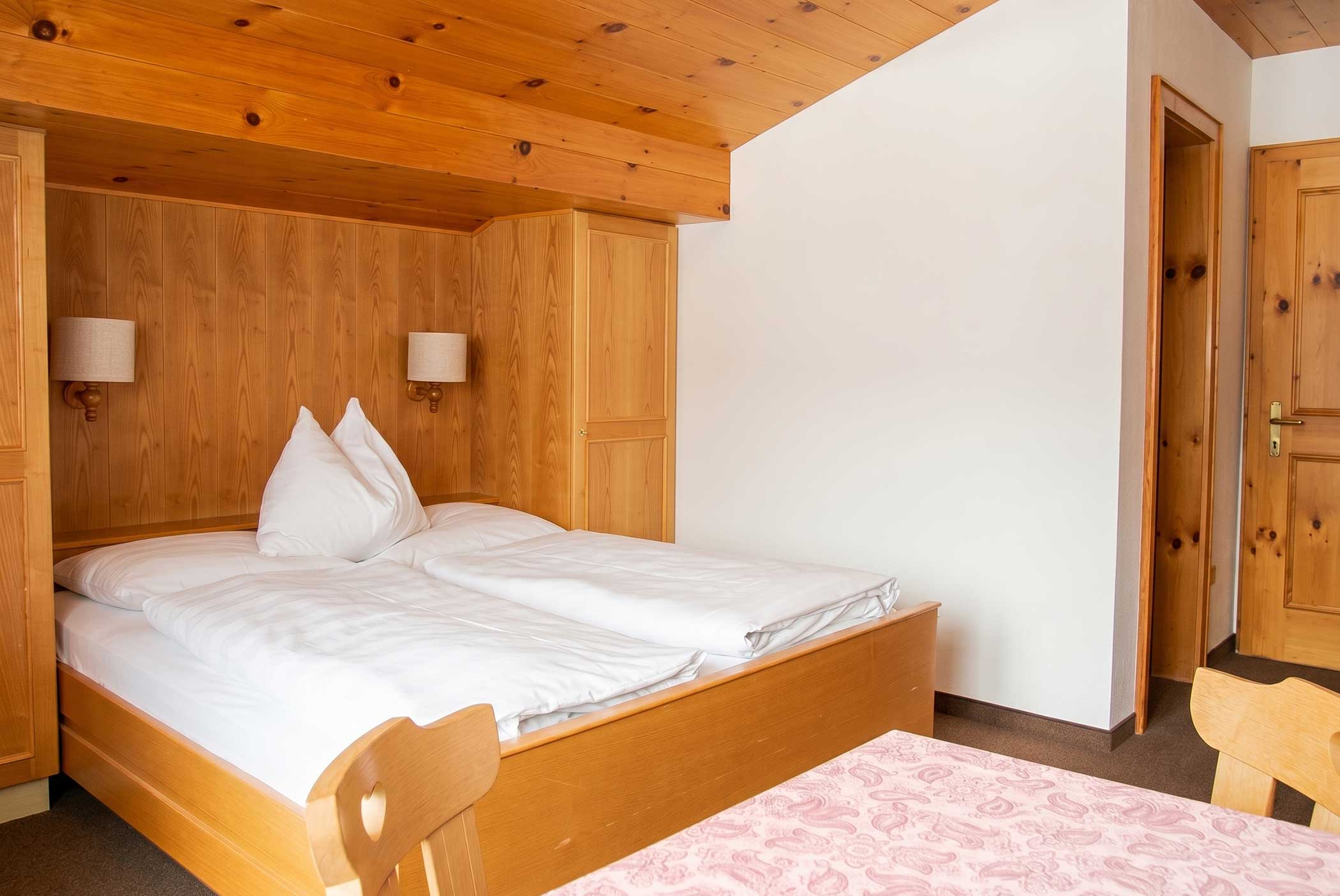 Gemütliches Schlafzimmer mit Holzmöbeln, einem Doppelbett, weißem Bettzeug und einer rosa Tischdecke auf dem Tisch. - Hotel Mooserhof