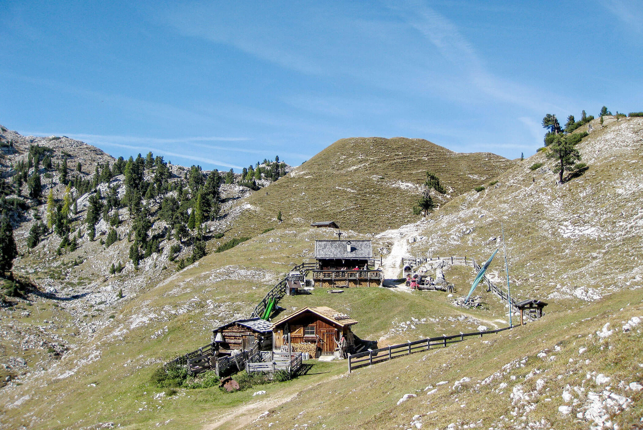 Holzhütten an einem grasbewachsenen Hang mit felsigen Bergen und Bäumen unter einem klaren blauen Himmel. - Hotel Mooserhof