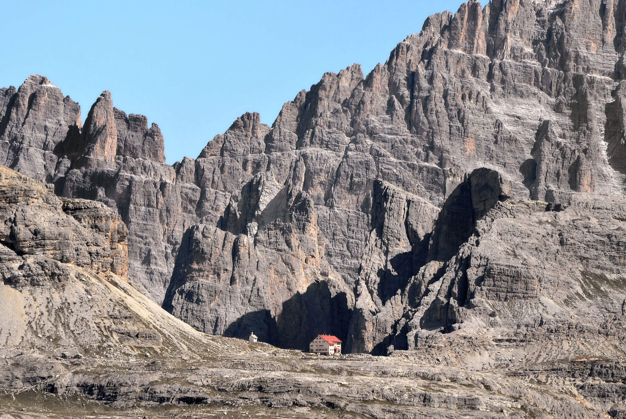 Ein kleines Haus mit rotem Dach steht am Fuße hoher, zerklüfteter Berge unter einem klaren blauen Himmel. - Hotel Mooserhof