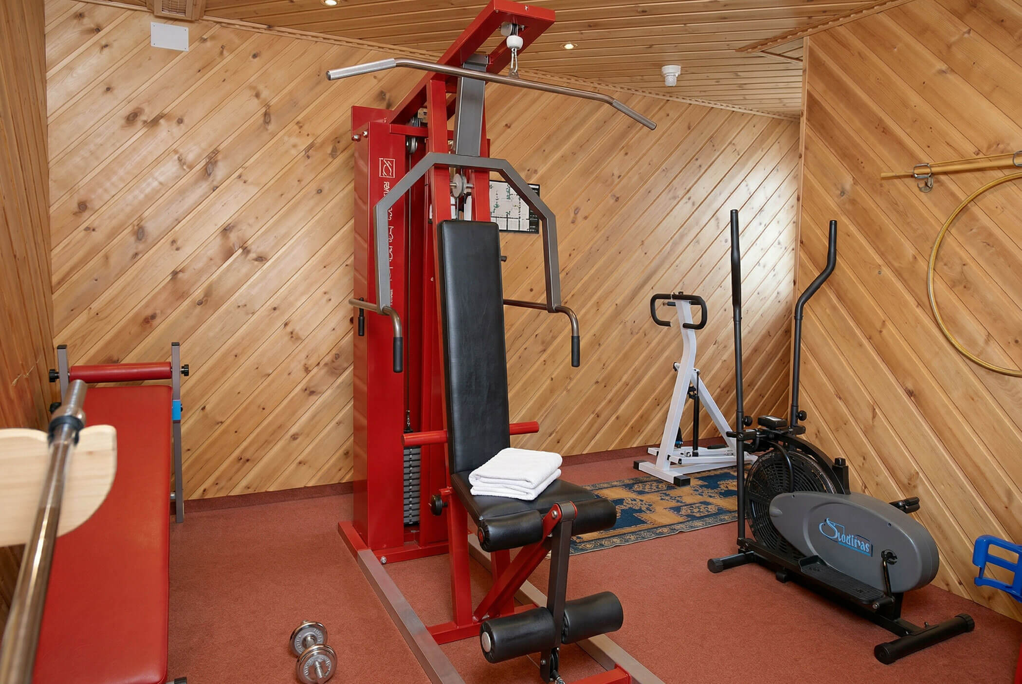 Kleiner Fitnessraum mit Holzwänden, roter Kraftmaschine, stationären Fahrrädern, Bank und gefalteten Handtüchern. - Hotel Mooserhof