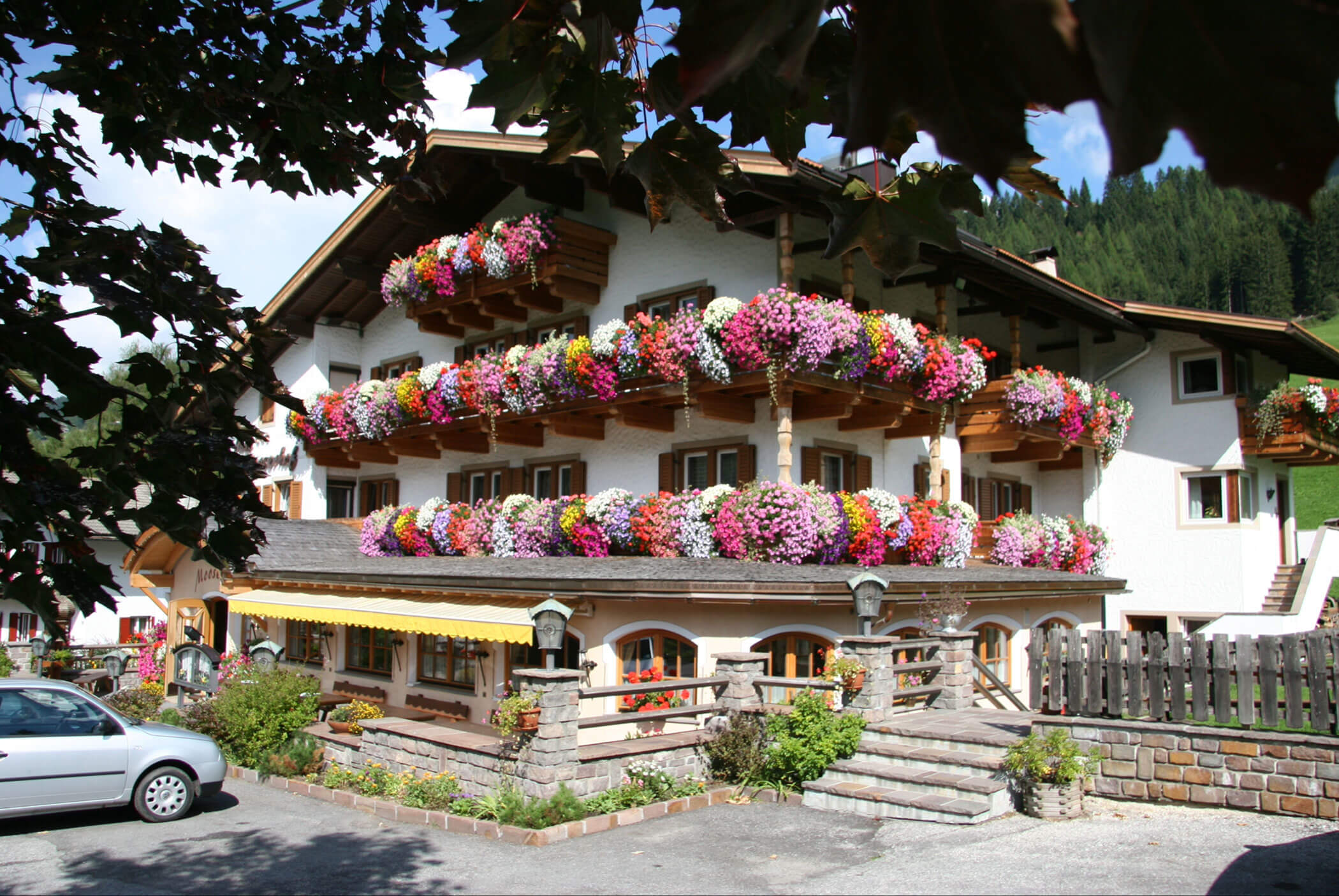 Alpenhaus mit Balkonen, die mit bunten Blumen bedeckt sind, umgeben von Grünflächen und einem geparkten Auto davor. - Hotel Mooserhof