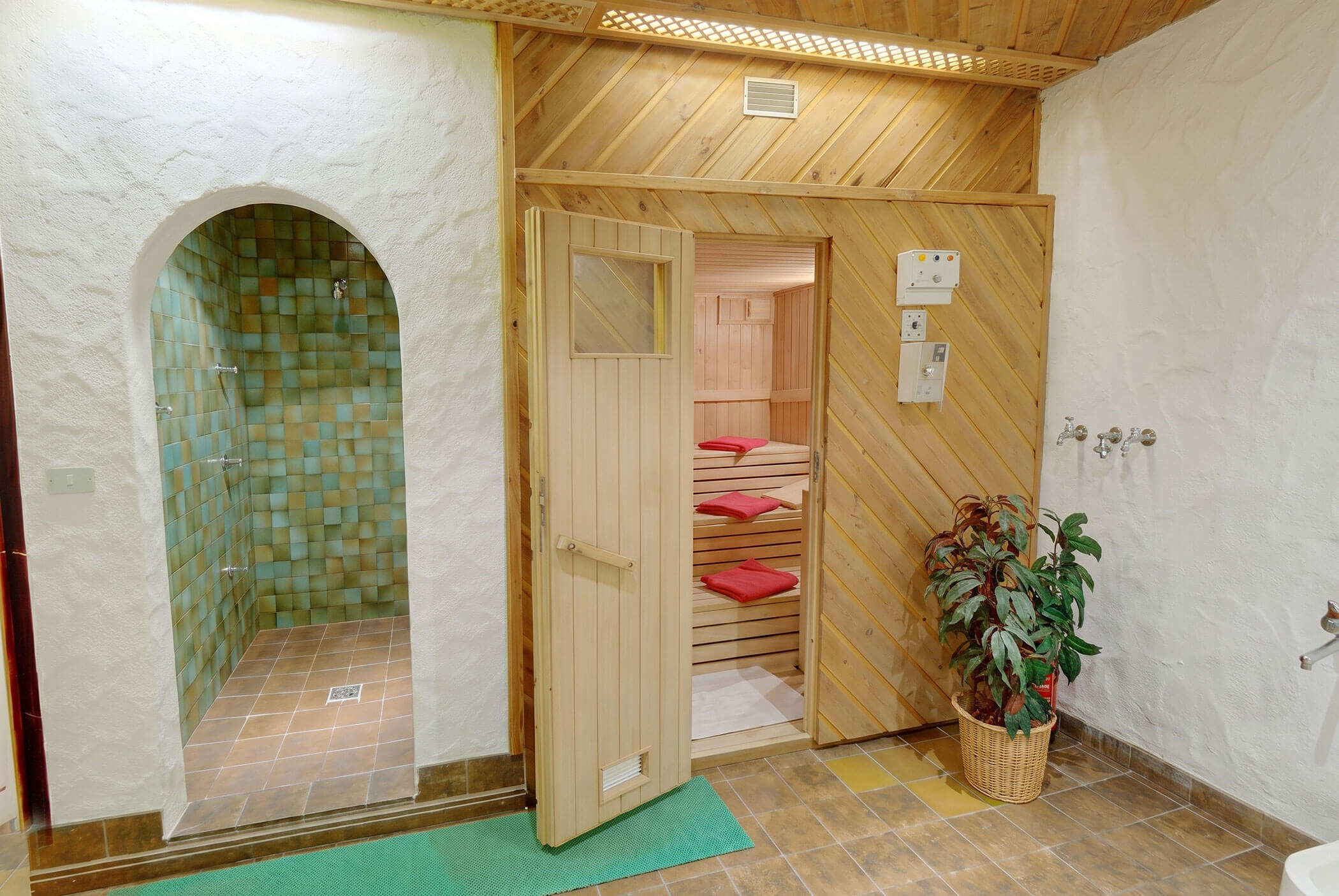 Eine Holzsauna mit roten Handtüchern, daneben ein gefliester Duschbereich und eine Topfpflanze auf dem Boden. - Hotel Mooserhof